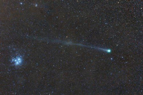 A comet with a bright green tail streaks across a star-filled night sky, near a cluster of stars.