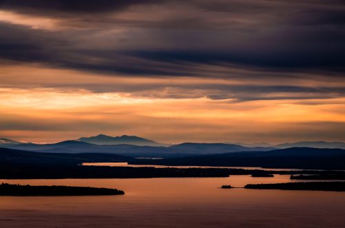 A serene landscape at sunset, featuring a lake surrounded by mountains and dramatic, colorful clouds in the sky.