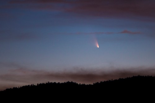 A comet streaks across a twilight sky above a dark silhouette of mountains.