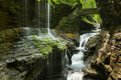 A serene landscape featuring cascading waterfalls flowing through rocky cliffs, with a stone bridge in the background.