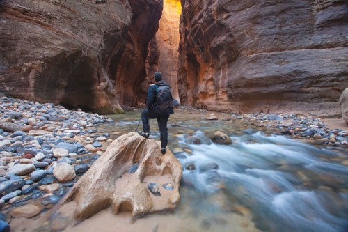 A person stands on a rock by a river in a narrow canyon, surrounded by towering stone walls and smooth pebbles.