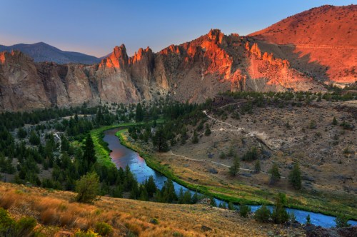 A scenic view of a river winding through rocky cliffs and green trees at sunset, with mountains in the background.