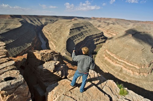 A child stands on a rocky ledge, photographing a vast canyon landscape with a river below.
