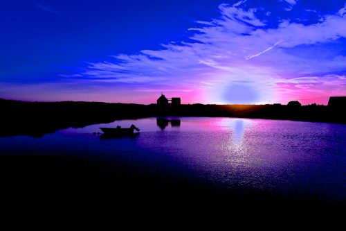 A serene sunset over a calm water body, with silhouettes of a boat and buildings against a vibrant sky.
