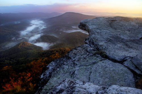 A rocky cliff overlooks a misty valley at sunrise, with rolling hills and a colorful sky in the background.