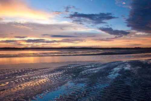A serene sunset over a calm water body, with colorful clouds reflecting on the surface and rippled sand in the foreground.