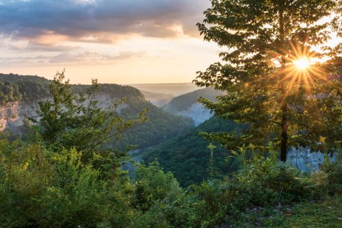Sunset over a lush green valley, with trees in the foreground and a golden sunburst peeking through the clouds.