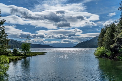 A serene lake surrounded by lush greenery and mountains, with fluffy clouds reflecting in the water under a bright sky.