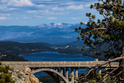 A scenic view of a bridge over a lake, surrounded by mountains and trees under a blue sky with clouds.