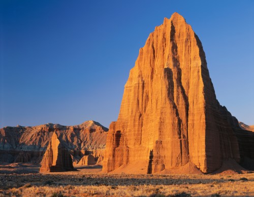 A towering rock formation bathed in warm sunlight, surrounded by rugged terrain and clear blue skies.