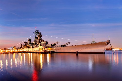 A battleship docked at sunset, reflecting lights on calm waters, with a clear sky in the background.