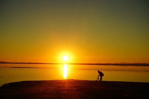 A silhouette of a person with a camera at sunset, reflecting golden light on calm water.