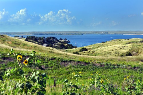 A scenic view of rolling green hills leading to a calm lake, dotted with sailboats under a blue sky with fluffy clouds.