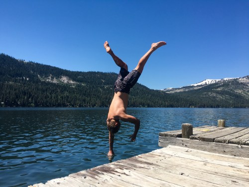 A boy performs a handstand on the edge of a dock over a lake, surrounded by mountains and clear blue skies.