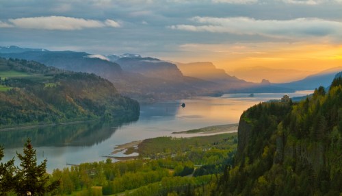 A scenic view of a river valley at sunset, surrounded by mountains and lush greenery. Soft clouds in the sky.