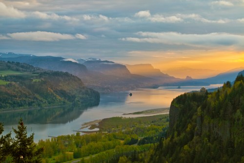 A serene landscape at sunset, featuring a river winding through lush green hills and distant mountains under a cloudy sky.