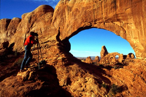 A photographer captures a stunning rock arch in a desert landscape under a clear blue sky.