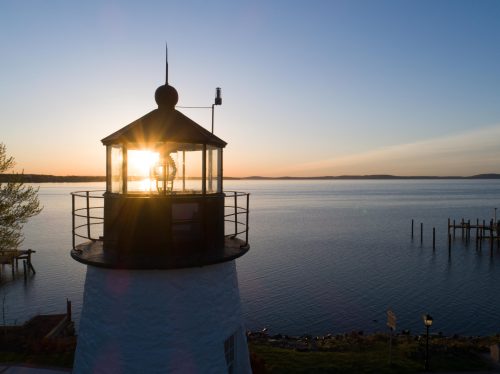 A lighthouse silhouette against a sunset over calm waters, with a pier in the background.