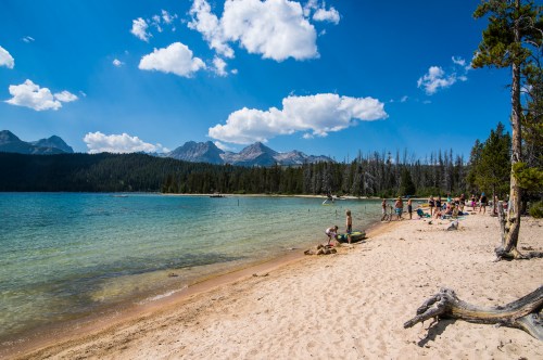 A sunny lakeside scene with people enjoying the beach, mountains in the background, and fluffy clouds in the blue sky.