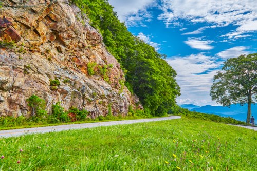 A scenic view of a rocky hillside with lush greenery, a winding road, and a clear blue sky in the background.