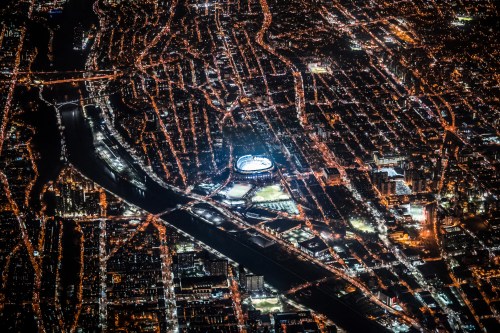 Aerial view of a city at night, featuring illuminated streets and a stadium near a river.