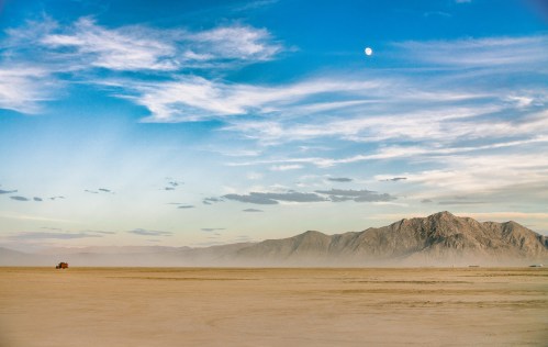 A vast desert landscape with mountains in the background, a clear blue sky, and a bright moon overhead.