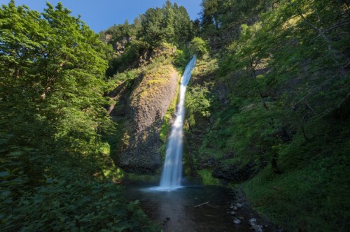 A tall waterfall cascades down rocky cliffs, surrounded by lush green trees and foliage under a clear blue sky.