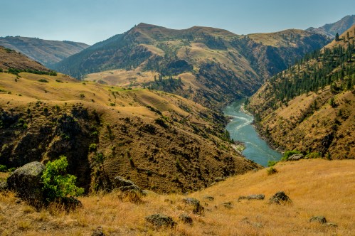 A winding river flows through a rugged canyon surrounded by rolling hills and sparse vegetation under a clear sky.