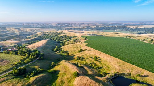 Aerial view of rolling hills and green fields under a clear blue sky, with a small pond visible in the landscape.