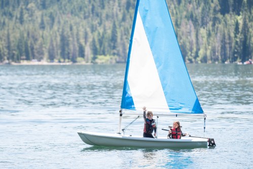 Two children in life jackets sailing on a small boat with a blue sail on a calm lake surrounded by trees.