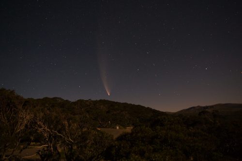 A comet streaks across a starry night sky above a dark, wooded landscape.