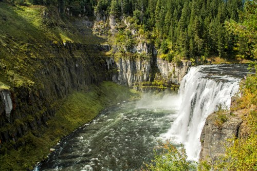 A stunning waterfall cascades into a river, surrounded by lush green cliffs and trees under a clear blue sky.