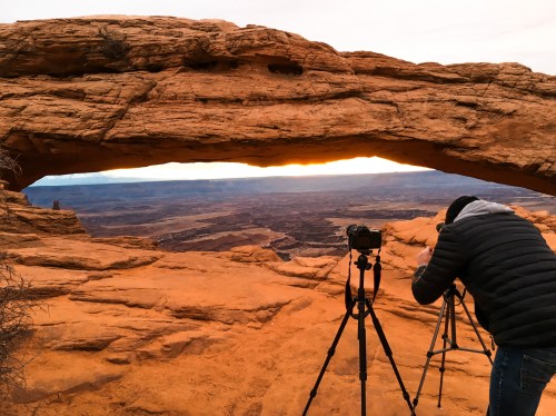 A photographer sets up a camera under a rock arch, capturing a sunset over a vast canyon landscape.