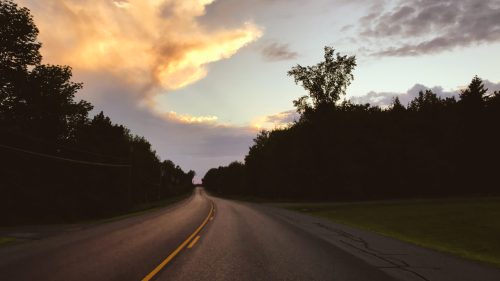 A winding road stretches into the distance under a colorful sunset sky, framed by trees on either side.