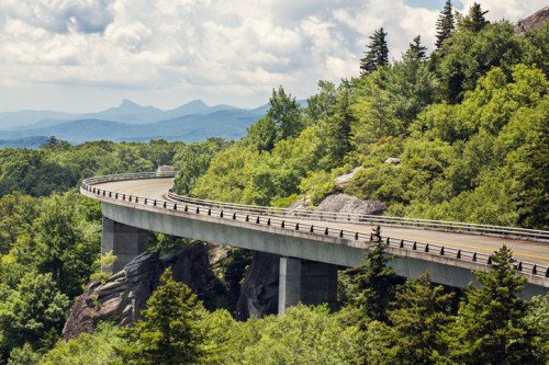A winding highway bridge curves through lush green mountains under a cloudy sky.