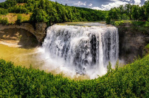 A stunning waterfall cascades over rocky cliffs, surrounded by lush greenery and a cloudy blue sky.