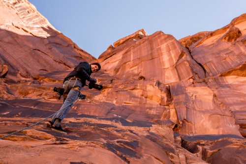 A climber ascends a rocky cliff, capturing photos with a camera, surrounded by towering red rock formations.