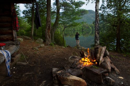A campfire in the foreground with a person standing by a lake, surrounded by trees and mountains in the background.