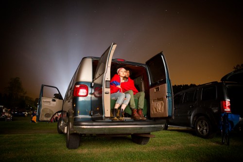 A couple sits in the open back of a van, gazing at the night sky filled with stars.