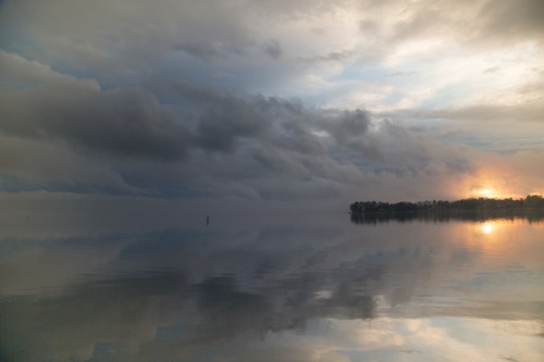 A serene lake at sunset, with calm waters reflecting dark clouds and a hint of orange light on the horizon.