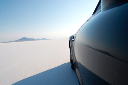 A sleek car drives on a vast, white salt flat with distant mountains under a clear blue sky.