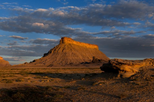 A dramatic desert landscape featuring a prominent rock formation under a blue sky with scattered clouds.