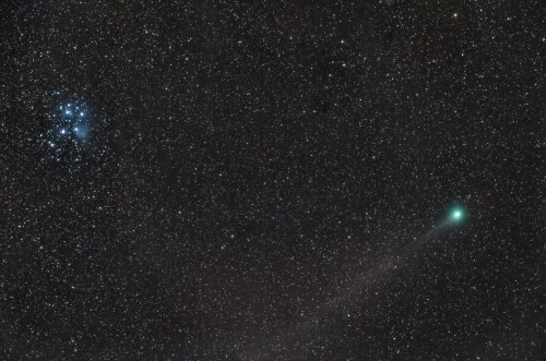 A bright green comet trails through a starry night sky, near a cluster of stars.