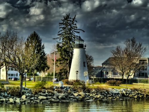 A white lighthouse stands by the water, surrounded by trees and buildings, under a dramatic cloudy sky.