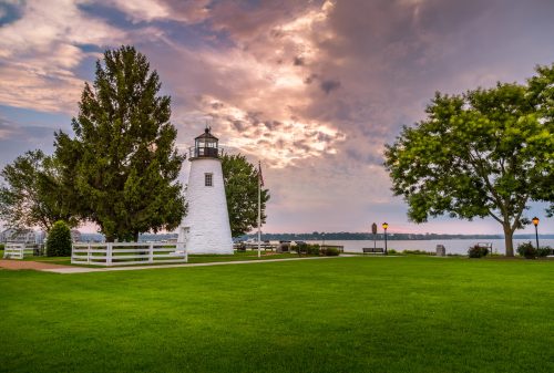 A white lighthouse stands on a grassy area, surrounded by trees, with a colorful sky and water in the background.