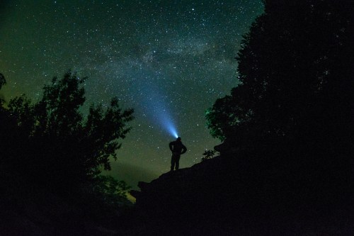 A silhouette of a person with a flashlight standing on a rock, gazing at a starry night sky.