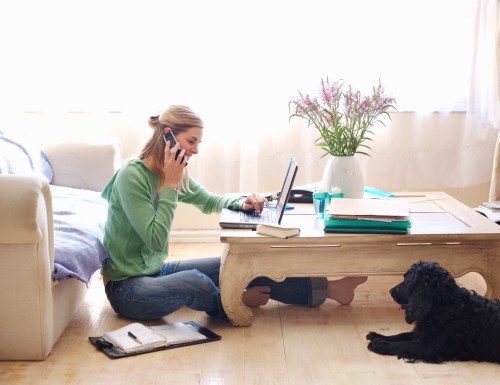 A woman sits on the floor, talking on the phone while working on a laptop, with a dog nearby and flowers in a vase.