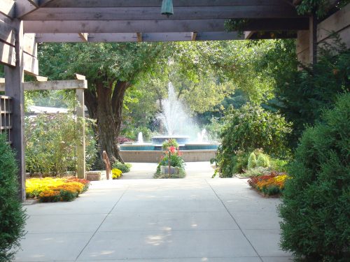 A serene garden pathway leads to a fountain, surrounded by lush greenery and colorful flowers.
