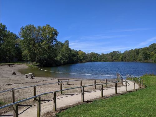 A serene lake surrounded by trees, with a sandy shore and a wooden railing leading to the water under a clear blue sky.