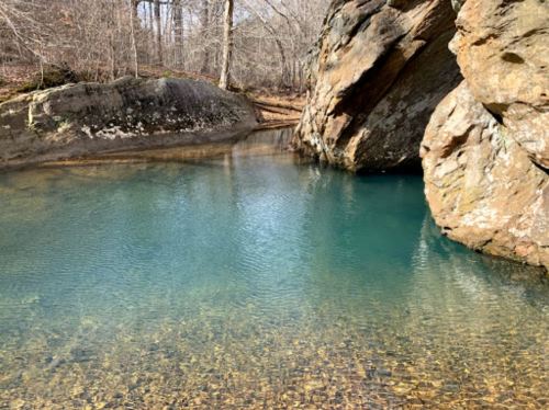 A serene, clear blue pool of water surrounded by rocky terrain and trees in a tranquil natural setting.